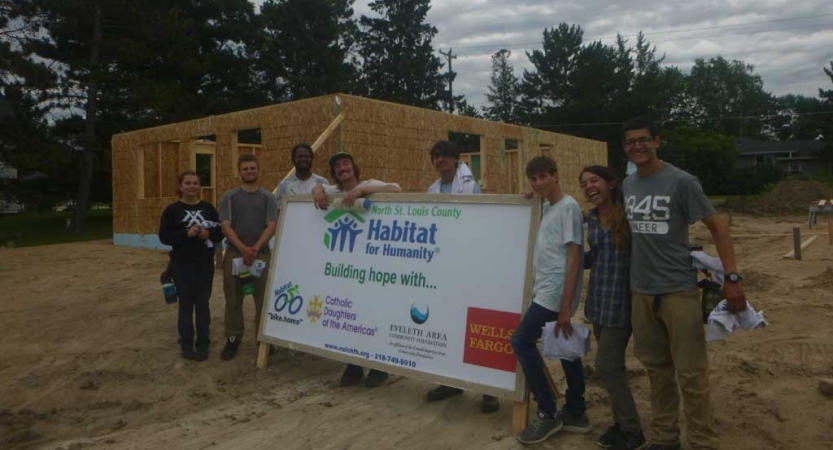 A group of young people pose for a photo around a habitat for humanity sign. Behind them is the frame of a house.